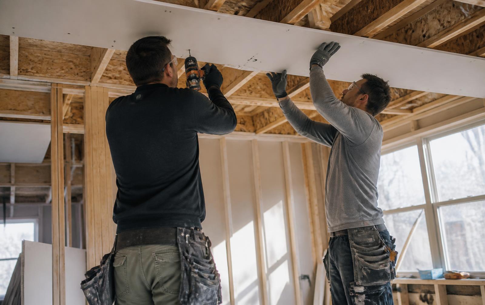 Two drywall installers fastening a gypsum panel to ceiling framing in a suburban home